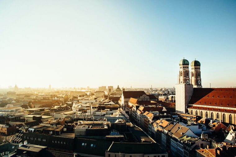 Blick über ein dicht bebautes Häusermeer einer deutschen Großstadt in warmem Abendlicht, die Türme der Münchner Frauenkirche am Horizont.