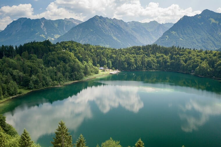 Der Freibergsee bei Oberstdorf im Sommer: klares, grün schimmerndes Wasser vor Alpenkulisse, am bewaldeten Ufer ein kleines Strandbad.