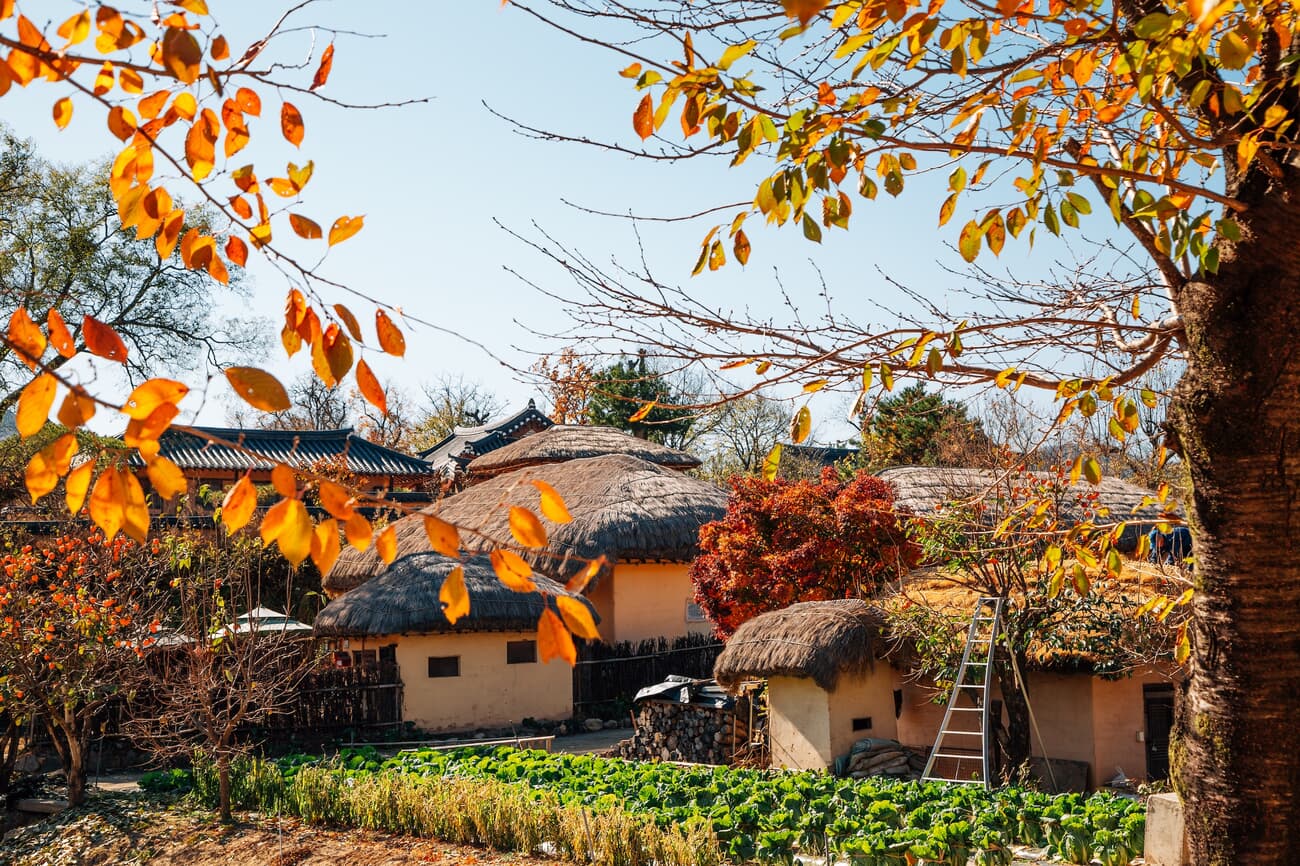 A traditional village with thatched-roof houses, surrounded by autumn foliage and a garden with gree