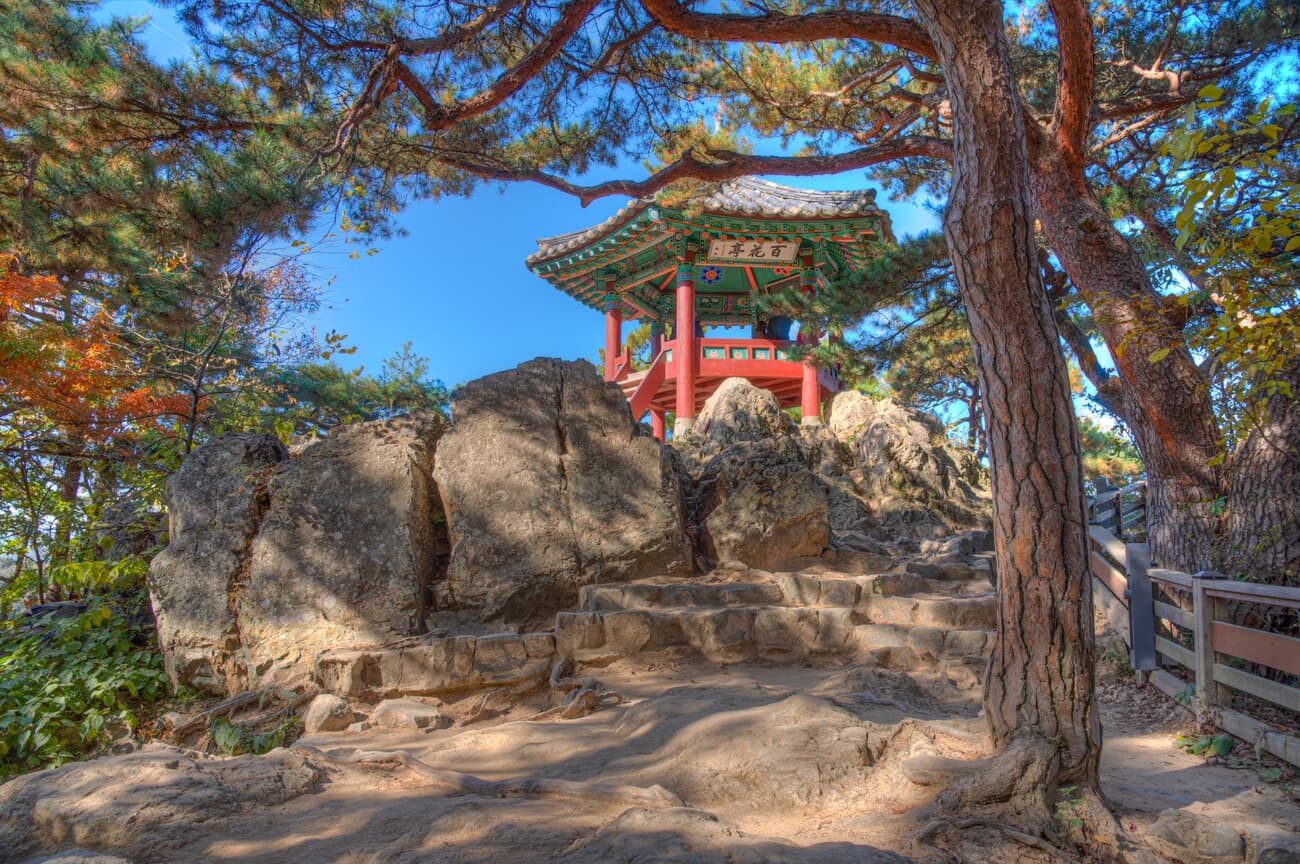 A traditional Korean pavilion nestled on a rocky outcrop surrounded by pine trees under a clear blue