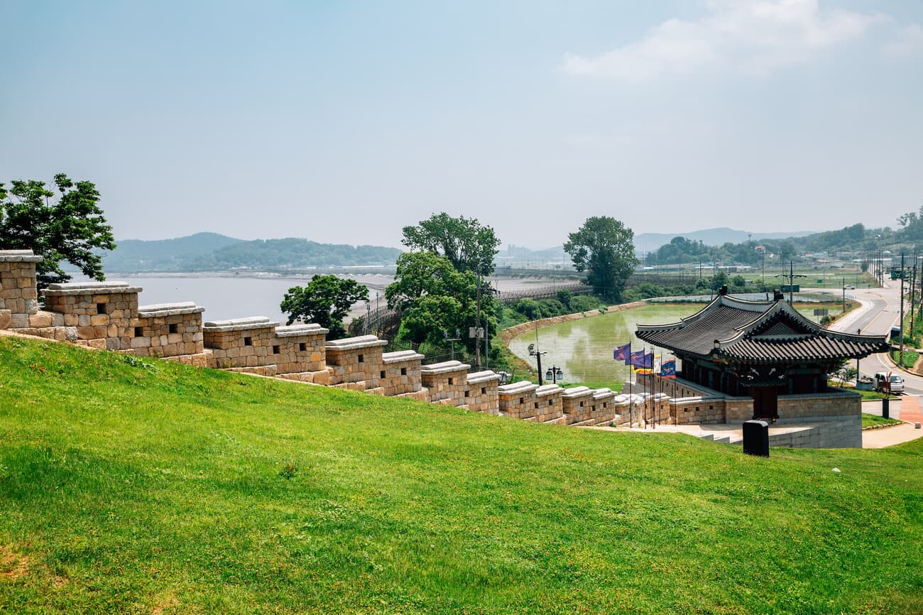 A traditional Korean pavilion next to a stone wall on a grassy hillside, with a river in the backgro