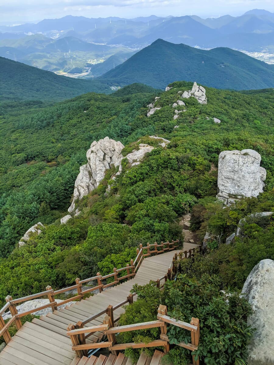 A wooden staircase winding down a lush mountain slope with large rocks and a panoramic view of dista