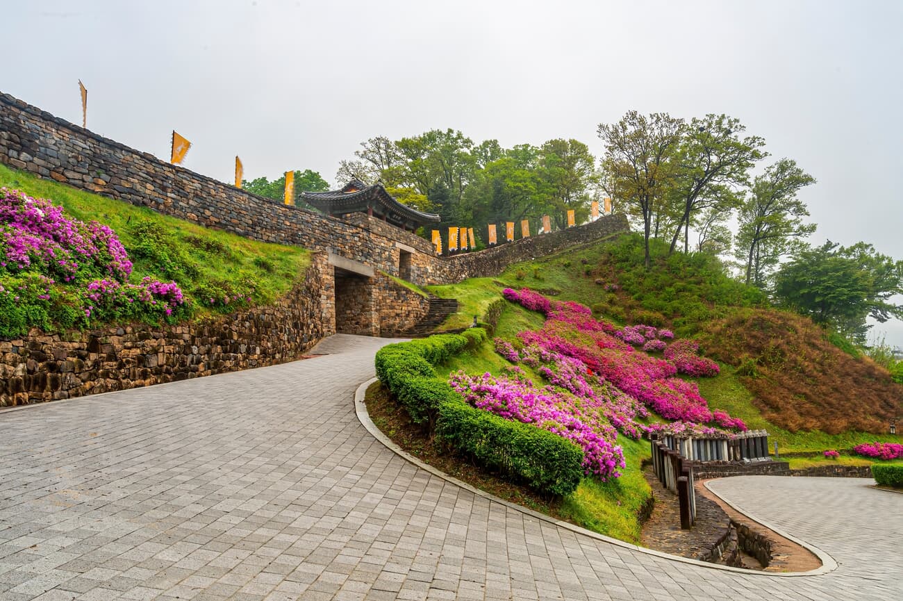 A winding cobblestone pathway leads to an ancient stone gateway adorned with traditional flags, surr