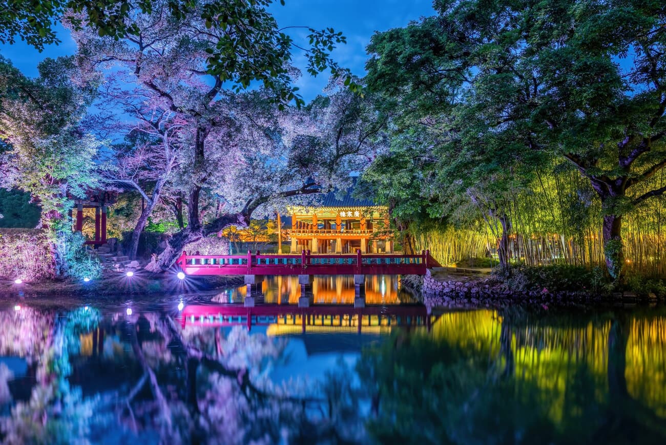 A traditional Asian pavilion illuminated at night, with a reflection in a tranquil pond, surrounded 