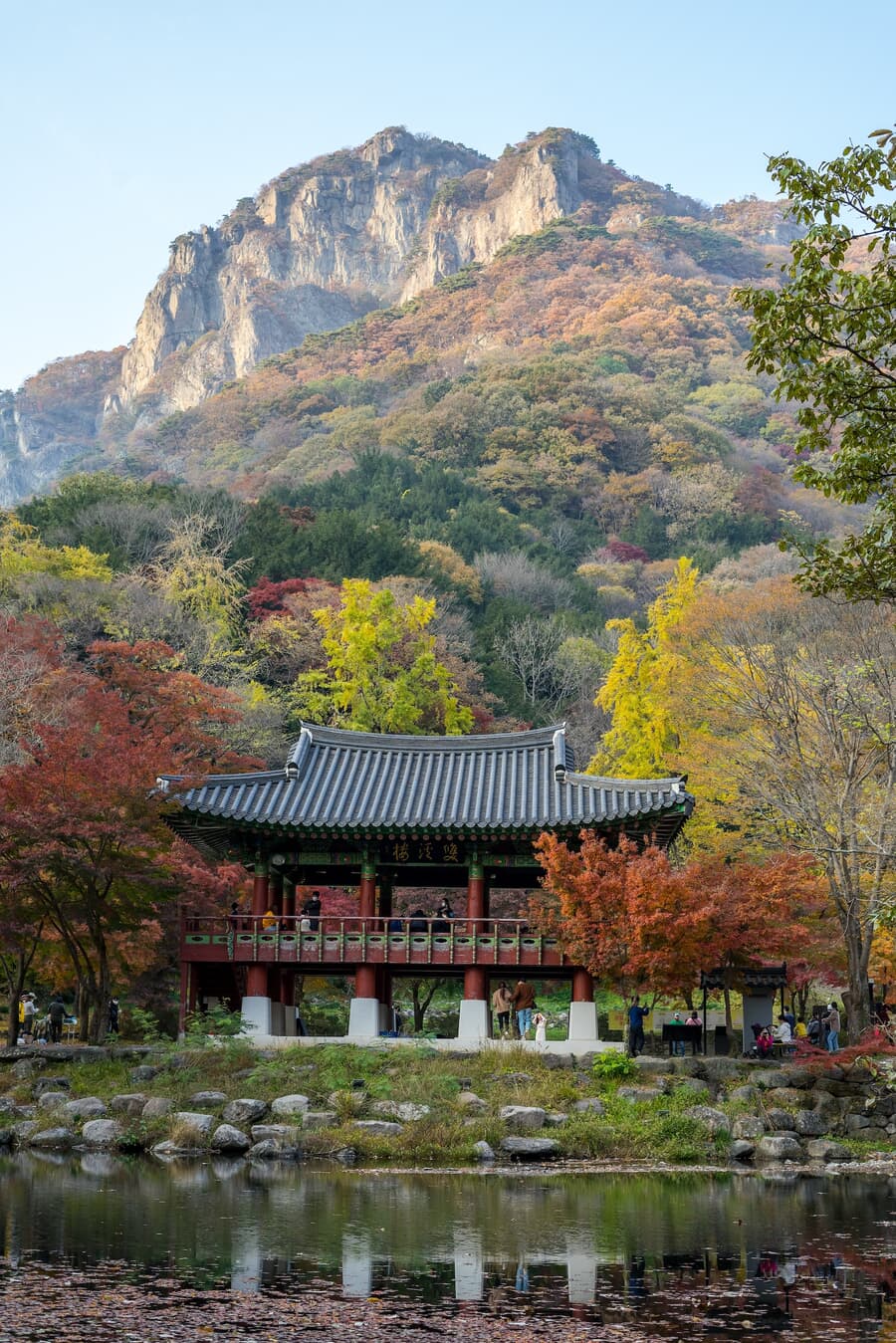 A traditional Korean pavilion by a tranquil pond with autumn-colored trees in the foreground and a r