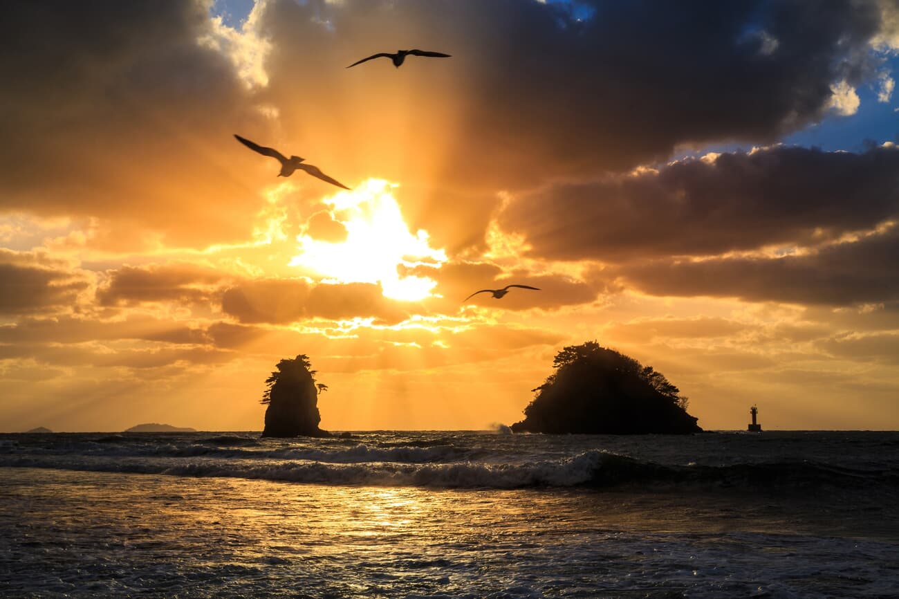 Sunset over the ocean with rays of sunlight piercing through clouds, highlighting rock formations an