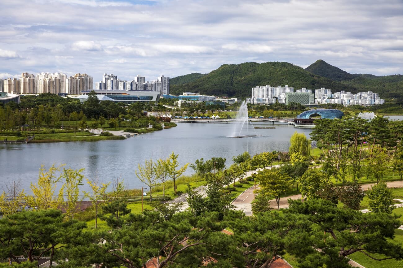 A scenic view of a city park with a large fountain in a lake, surrounded by lush greenery, walking p