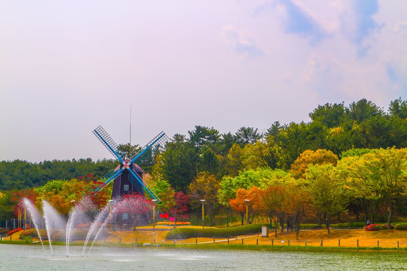 A traditional Dutch windmill beside a lake with multiple fountains, surrounded by colorful autumn tr