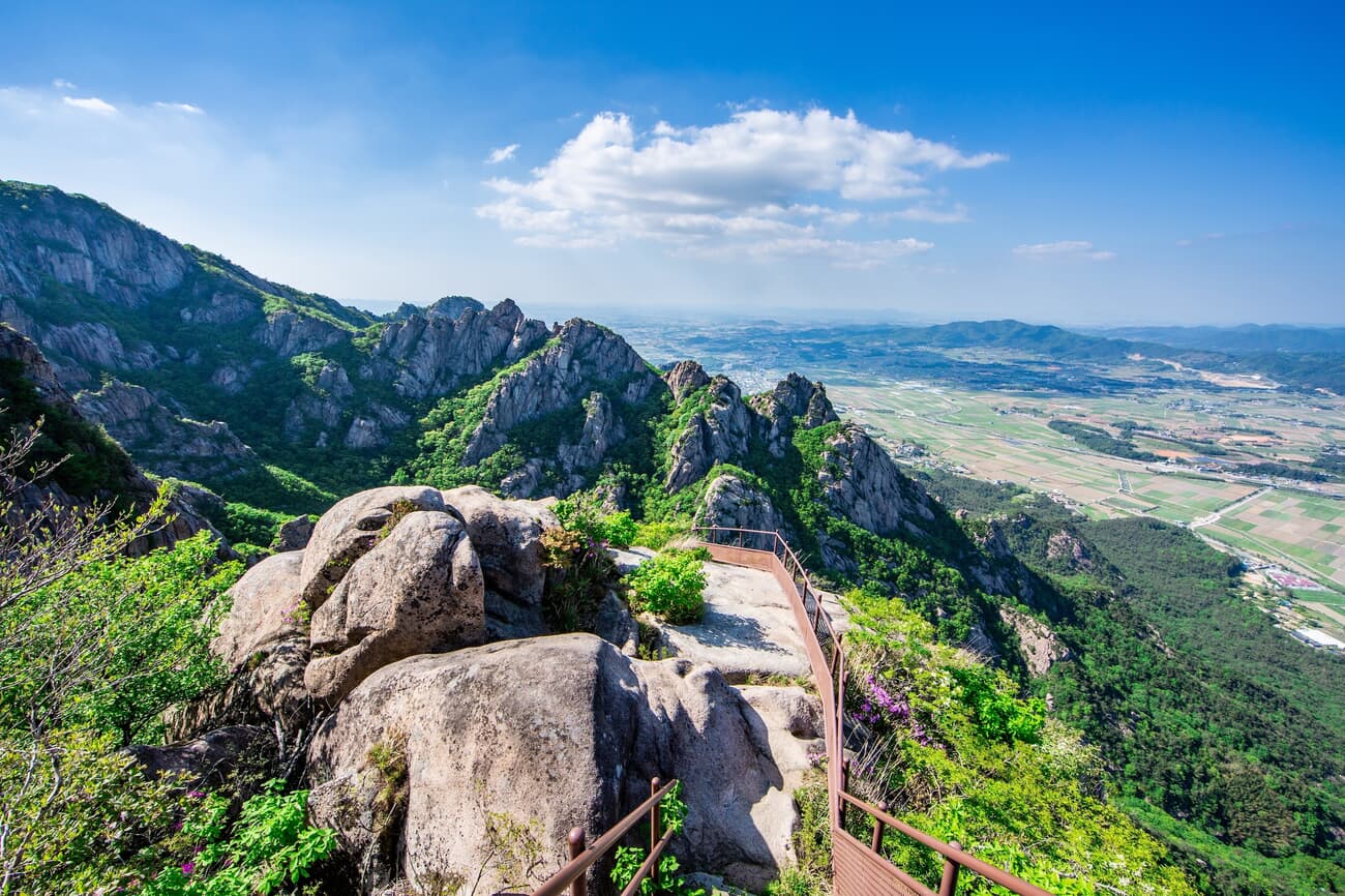 A scenic view from a mountain peak with a metal railing, overlooking a rugged landscape with sharp c