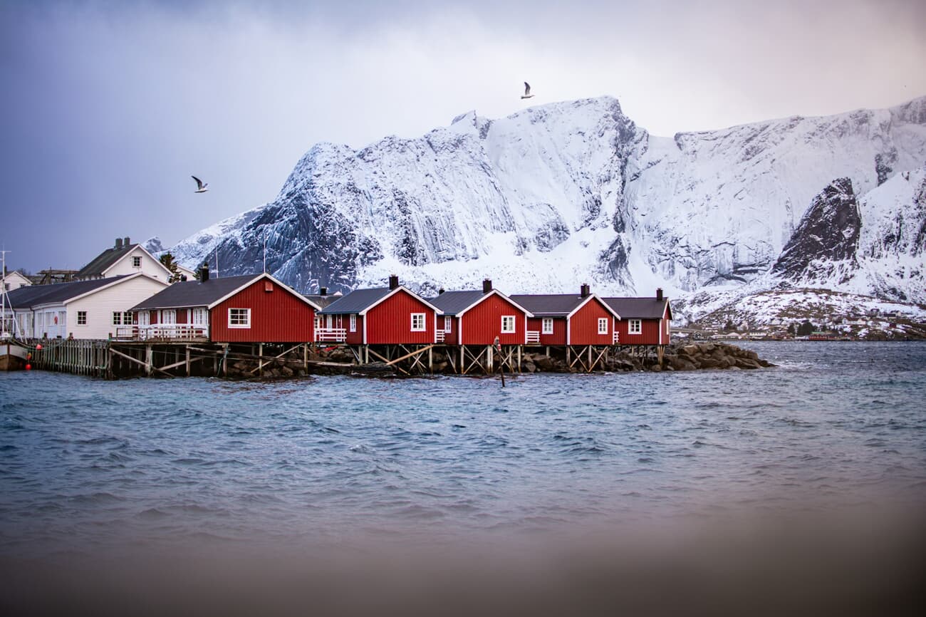 Red fishing cabins sit on the water beneath snow-covered peaks in Norway's Lofoten Islands