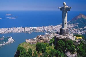 Aerial shot of Christ the Redeemer statue in Rio de Janeiro, Brazil, with city, bay, mountains, and clear skies in view.