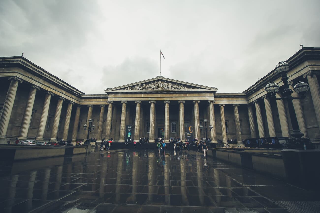 A wide-angle view of the British Museum in London on an overcast day, with visitors walking in front of the building's grand columned facade.