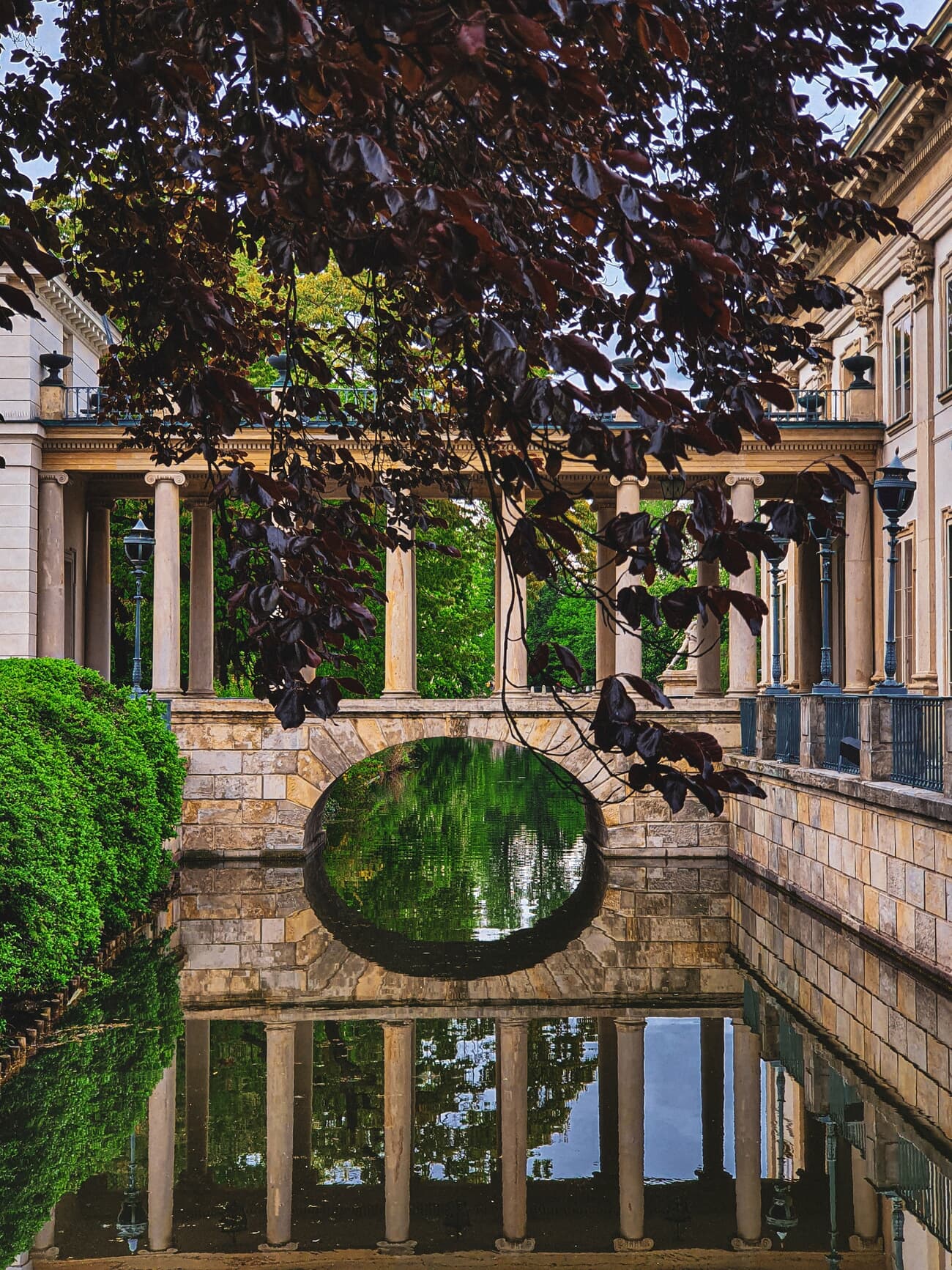 A serene view of ลazienki Park with a classical architecture building, a calm pond reflecting the structure, and lush greenery surrounding the area.