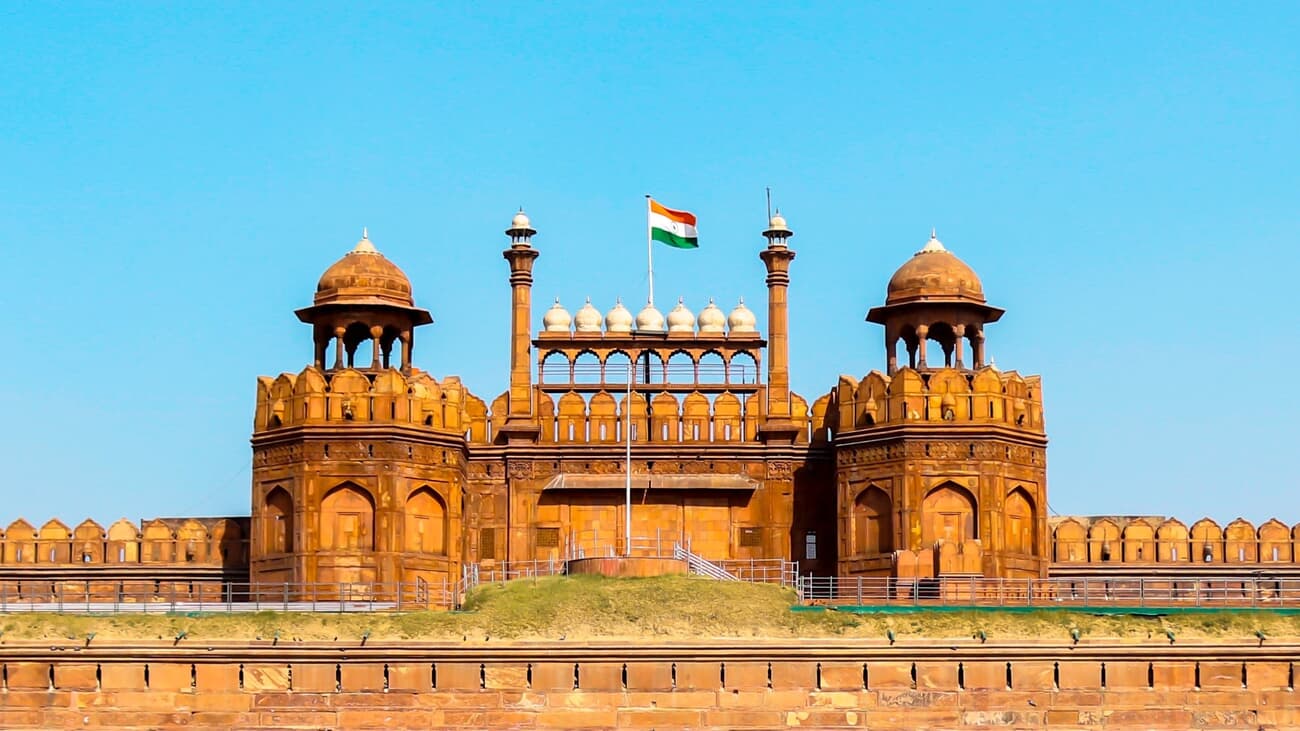 A photo depicts the Red Fort's red sandstone architecture, two domed towers, and the Indian flag under a clear blue sky in Delhi.