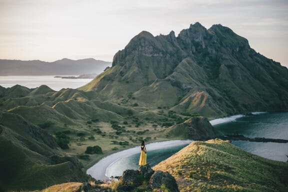 A woman in a yellow dress stands atop a hill on Padar Island in Indonesia.