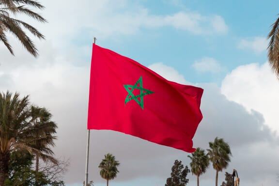 Flag of Morocco waving, with blue sky and clouds in the background and palm trees on the sides