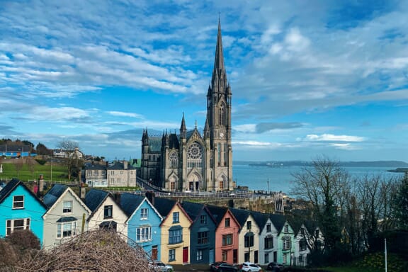 A dark grey church stands stall behind colorful residential buildings in Cobh, Ireland.