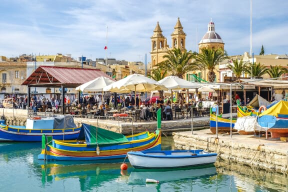Colorful fishing boats sit in the water next to land in Marsaxlokk, Malta.