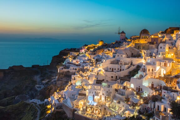 White buildings on a hillside are lit up at dusk in Oia, Greece.