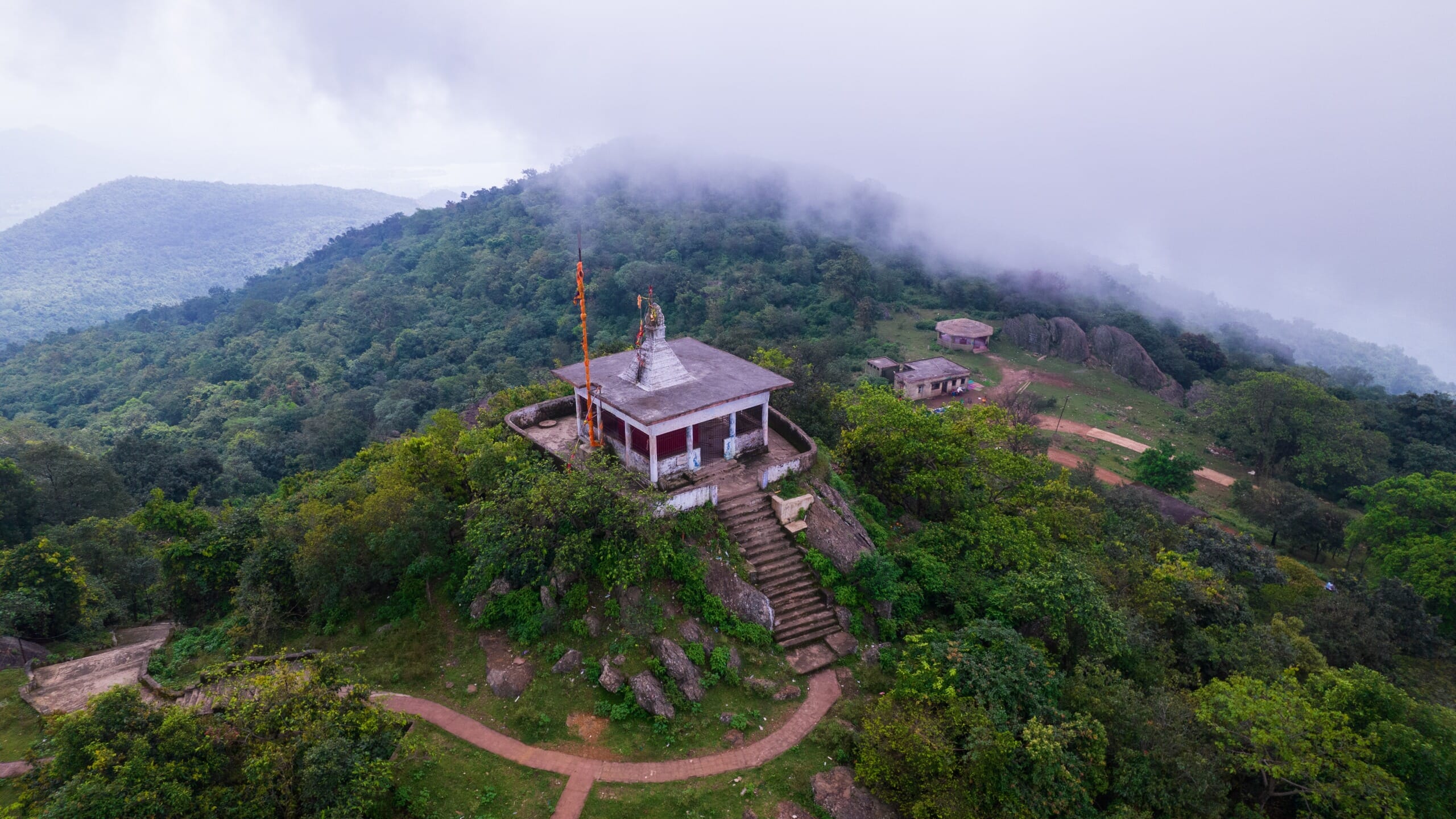 An aerial view of a small temple atop a green hill with low-lying clouds surrounding it.