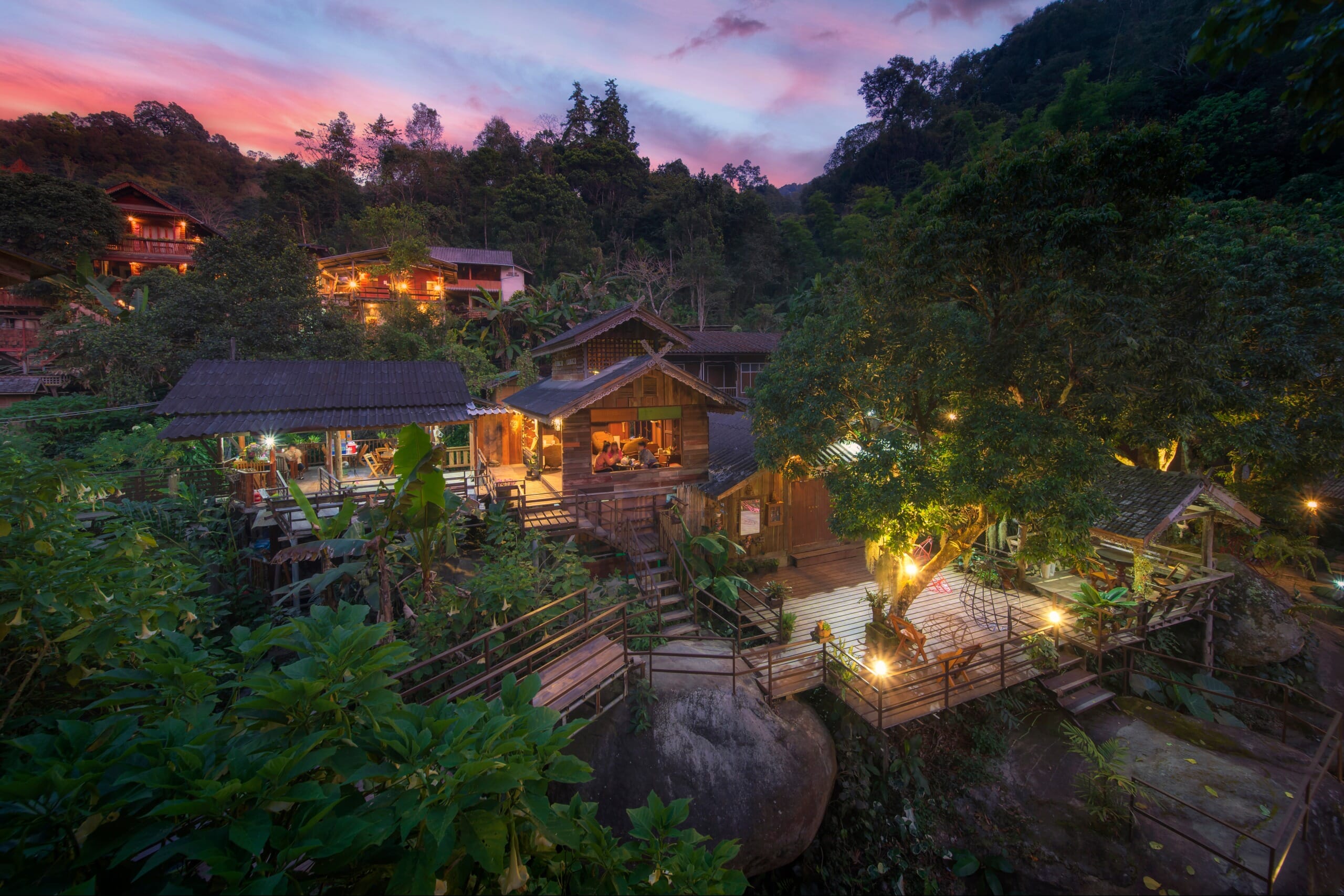 A serene mountain village at dusk with traditional wooden buildings surrounded by dense foliage, illuminated by warm lights against a twilight sky.
