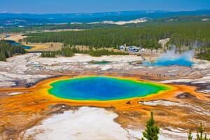 A vivid aerial view of the Grand Prismatic Spring in Yellowstone National Park with its bright colors ranging from blue to orange, tourists on a boardwalk, surrounding water formations, and a forested background.