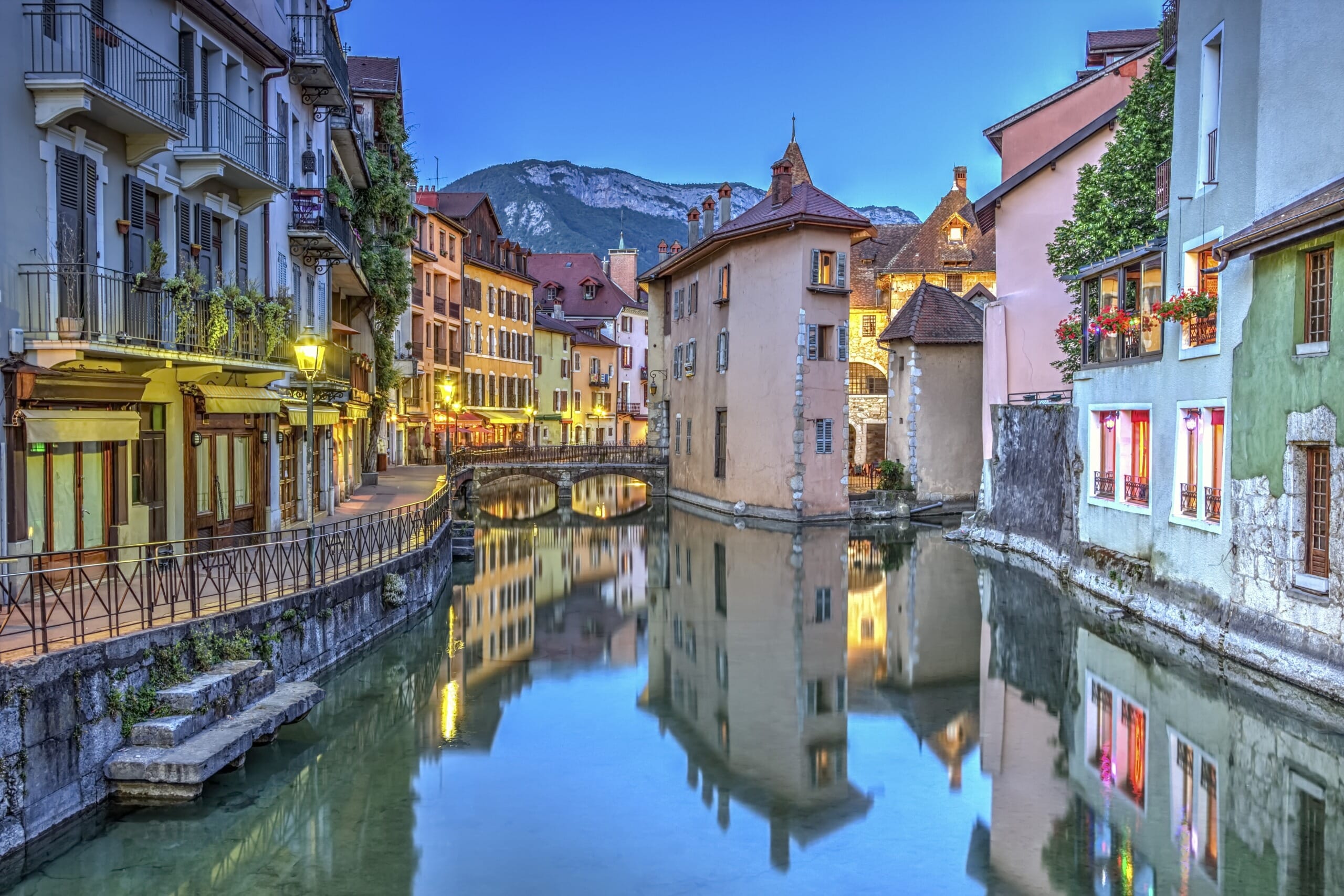 A picturesque twilight view of pastel-colored buildings lining a calm canal, with reflection on the water surface and a backdrop of mountains.