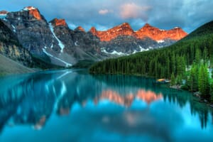 A serene scene of Moraine Lake in Banff National Park during twilight with sunlit mountain peaks reflected in the tranquil turquoise waters, surrounded by dense evergreen forests.
