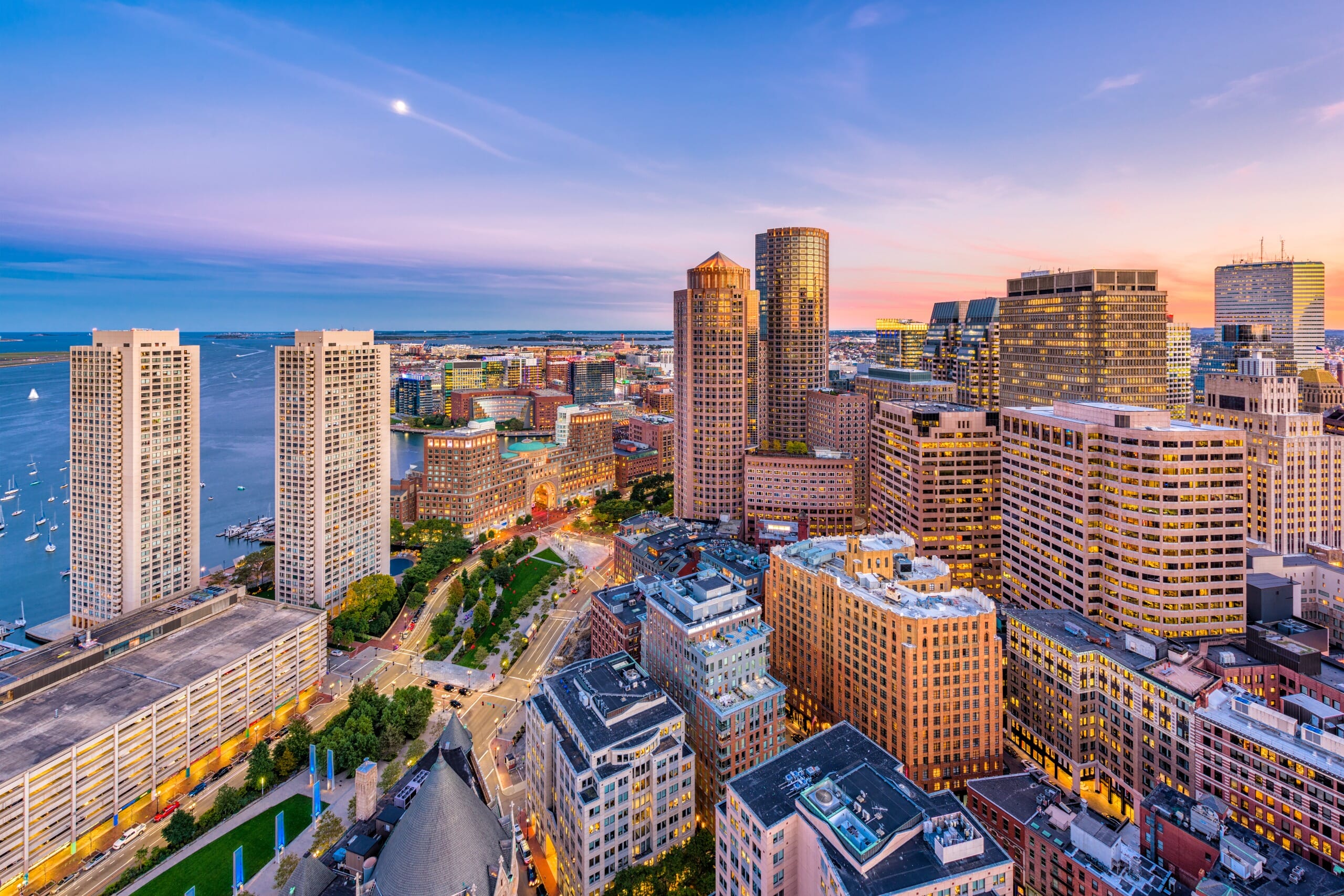 Aerial view of a densely populated city at dusk with skyscrapers, a waterfront, and a colorful sky.
