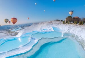 Hot air balloons floating above the terraced mineral pools of Pamukkale, Turkey, under a clear blue sky.