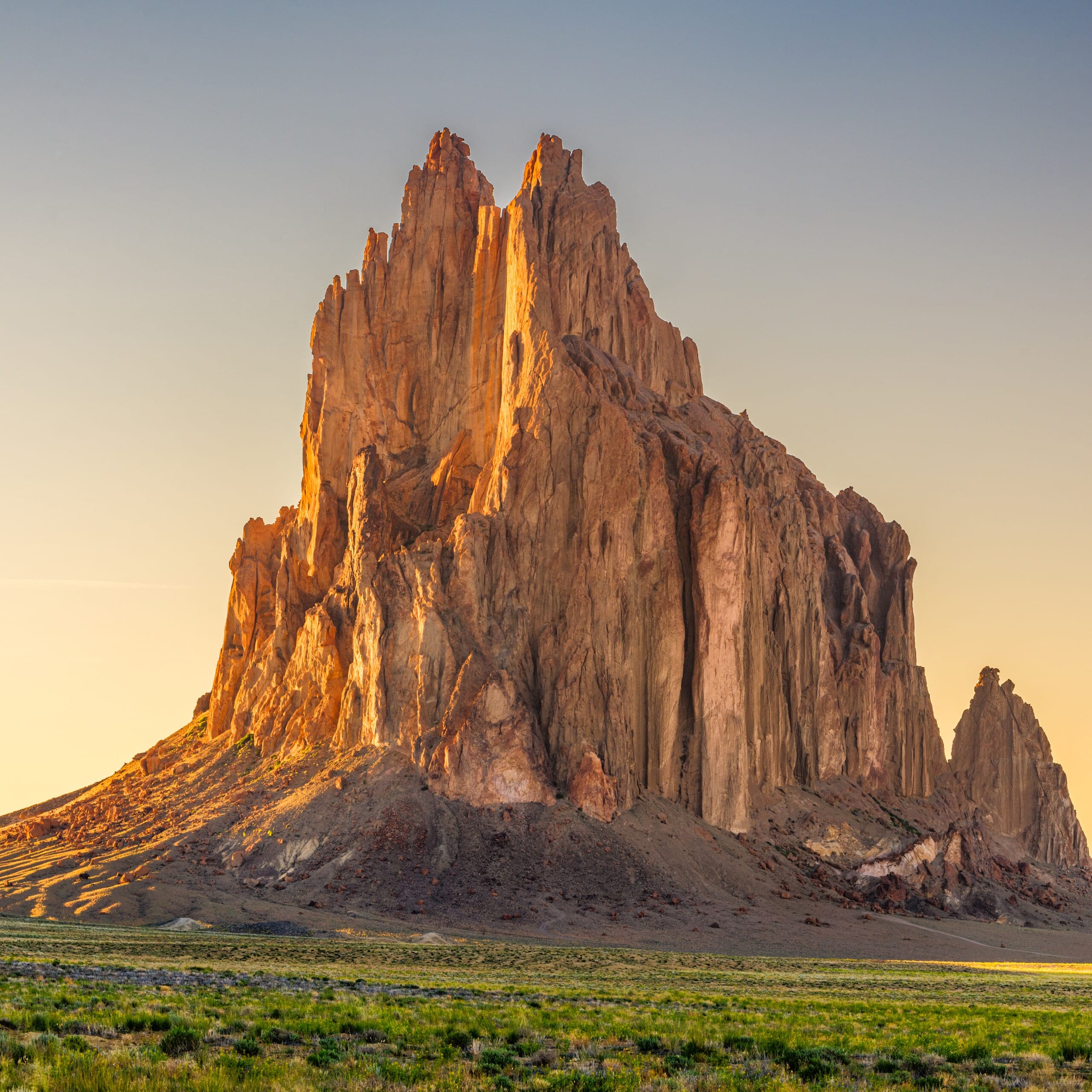 An imposing rock formation bathed in warm sunlight during golden hour, with sharp peaks rising against a clear sky and a foreground of green vegetation.