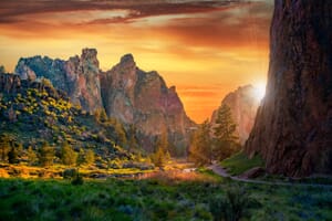 Sunset in a rugged mountainous landscape with vivid orange sky, the sun peeking through a narrow opening between towering cliffs, illuminating the foliage and a winding path in the valley.