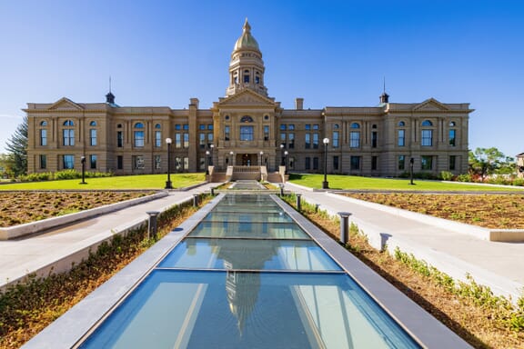 A majestic historical building with a central dome and clock, flanked by two symmetrical wings with a clear blue sky in the background. In the foreground, there is a reflecting pool leading up to the building
