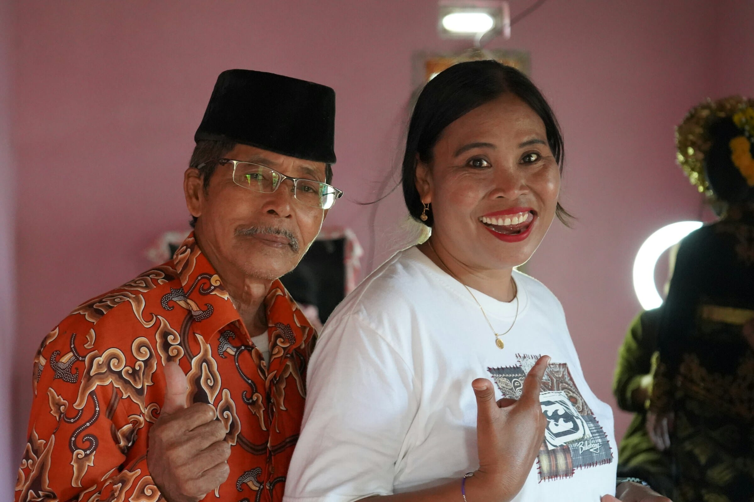 An older man wearing a black peci cap and orange batik shirt stands smiling beside a younger woman in a white shirt — a multigenerational portrait from Lombok, Indonesia, the country that topped the 2025 Global Flourishing Index.