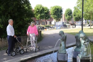 An older couple using walking aids strolls together along a park canal lined with sculptures — an image of later-life mobility and the years of healthy life expectancy that HALE measures.