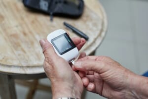 Older hands hold a blood-glucose meter with a test strip and a small blood droplet on the fingertip — the front-line diabetes screening tool that an estimated 252 million adults globally have never been tested with.